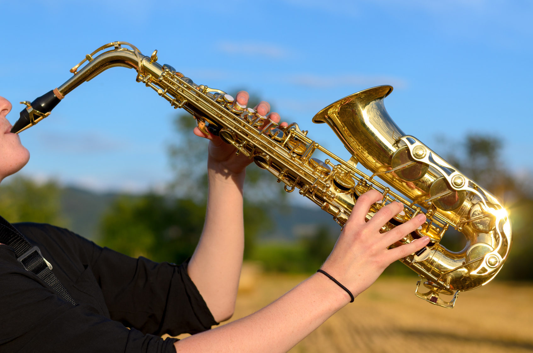 Side close up view of a young woman playing a tenor saxophone outdoors in the countryside raising the instrument in the air against a blue sky (Wird bei Klick vergrößert)